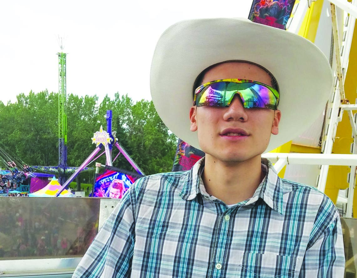 Young man in cowboy hat, sunglasses, blue plaid shirt and light blue jeans sitting on a ferris wheel