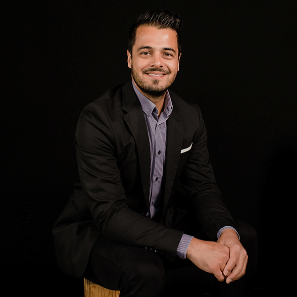 Raja Mita sits on a stool against a black background smiling. He is wearing a suit and has short hair, and a goatee