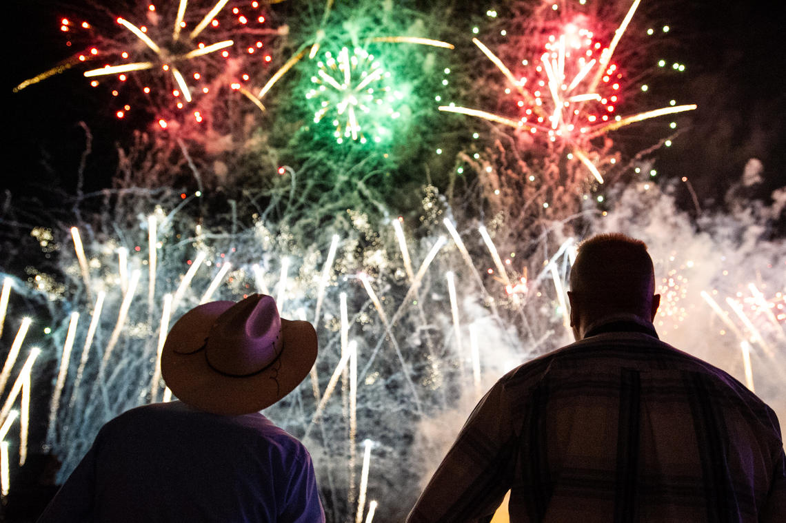 Two men stand silhouetted in front of red, green and white fireworks. One man wears a cowboy hat.