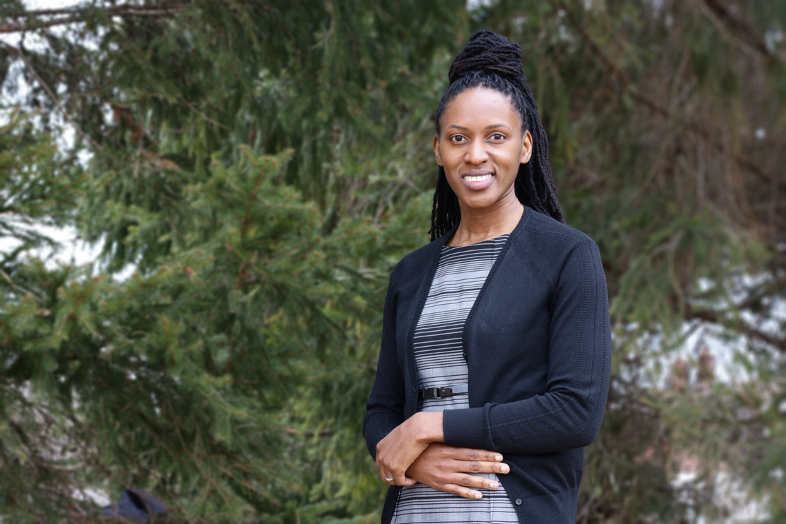 Patrina Duhaney stands smiling in front evergreen trees.