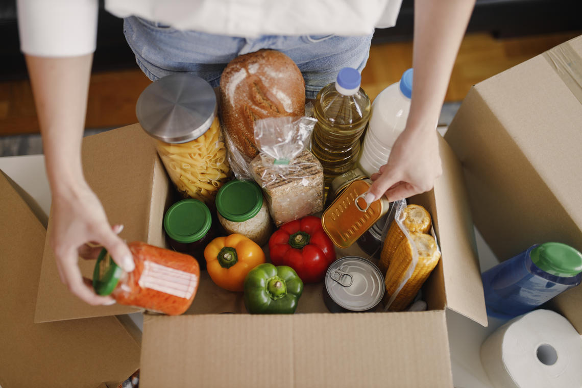 Person standing at a table with a box of food, interacting with a jar and a can of food inside the box.