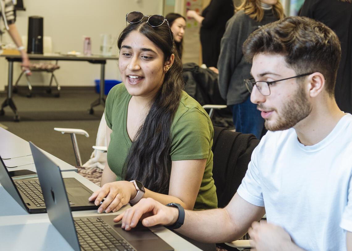 A young student with long hair and a young student with short hair look at a laptop and talk in a classroom.