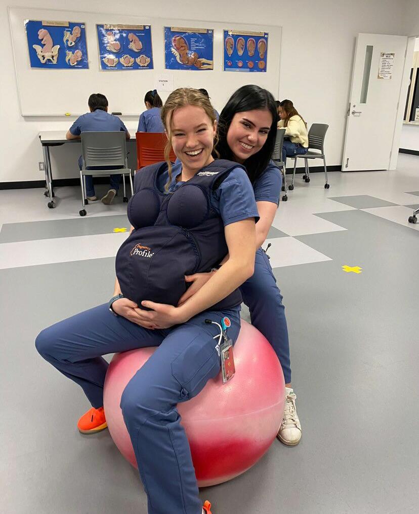 Holly sits on balance ball wearing pregnancy simulation belly while fellow nursing student practices aiding her