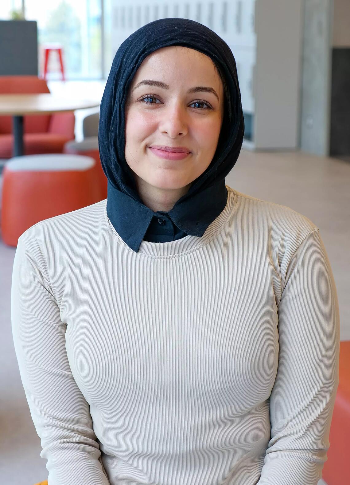 A woman wearing a hijab and collared shirt smiles at the camera