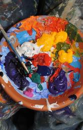 A brown bowl is filled with different colours of paint. A blue paintbrush rests in the bool, which is sitting on a paint splattered drop cloth.