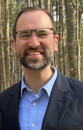 Matthew Makel, wearing glasses, a light blue shirt and a dark blue sport coat, stands smiling in front of trees.