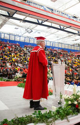 A man in a cap and gown speaks to a room full of graduates