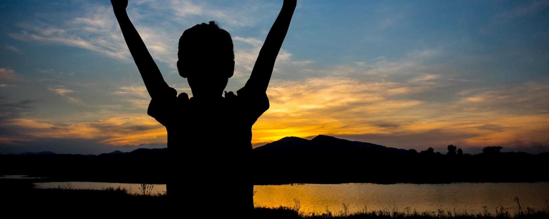 Child with raised arms facing a sunset