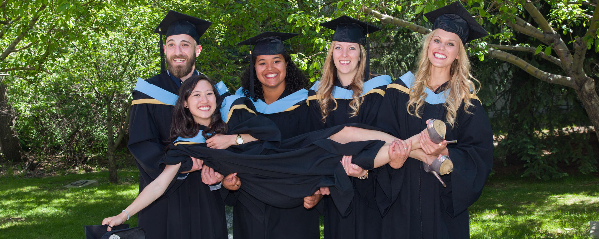 Five Werklund graduates wearing cap and gown pose for a photo on campus green space.